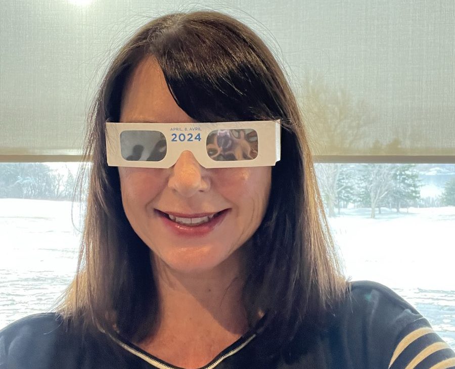A woman with long brown hair and bangs off to the side smiles and wears the free solar eclipse glasses that will be available at Fort Henry in Kingston.